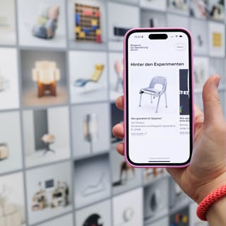 Hand hält Smartphone vor Wand mit Bildern, zeigt Stuhlbild auf Bildschirm.