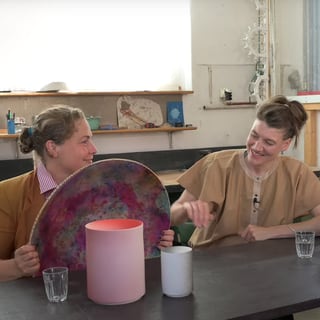 Zwei Frauen sitzen an einem Tisch mit bunten Objekten in einem Studio.