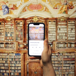 Person hält Smartphone vor Bibliothekswand mit Bücherregalen.