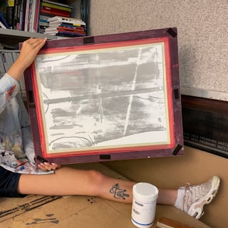 Person zeigt ein Siebdruckrahmen, umgeben von Kunstmaterialien in einem Atelier.