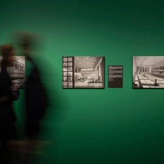 Zwei unscharfe Personen vor grüner Wand mit schwarz-weissen Fotos.