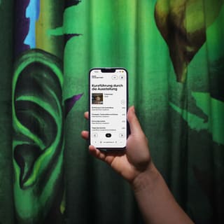 Hand hält ein Smartphone vor einem grünen Vorhang mit einem grossen Ohr.