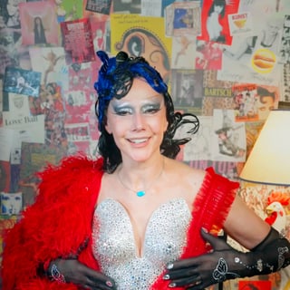 Person in glitzerndem Kostüm und roter Federboa, bunte Wand im Hintergrund.