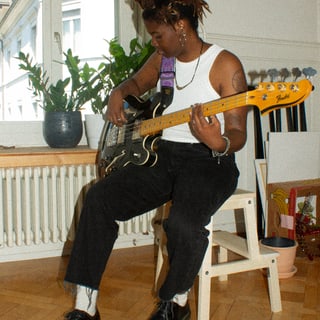 Person spielt E-Bass auf einem Stuhl sitzend in einem Zimmer mit Pflanzen im Hintergrund.