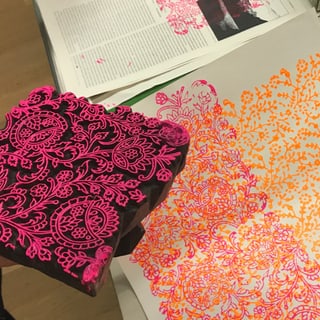 Holzblock mit floralem Muster neben bedrucktem Papier in Pink und Orange.",