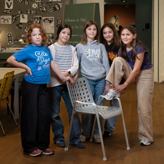 Fünf Kinder stehen in einem Raum neben einem Stuhl.