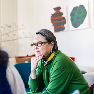 Person in grünem Pullover sitzt nachdenklich auf Sofa mit Kunstwand im Hintergrund.