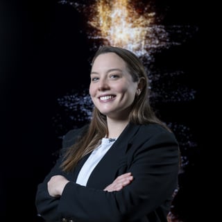 Frau in schwarzem Anzug vor digitalem Hintergrund, lächelt, Arme verschränkt.