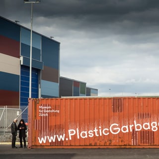 Lagercontainer mit Aufschrift 'PlasticGarbageProject.org' vor buntem Gebäude.