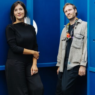 Zwei Personen stehen vor einer blauen Wand, lächelnd.