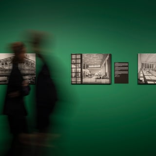 Zwei unscharfe Personen vor grüner Wand mit schwarz-weissen Fotos.