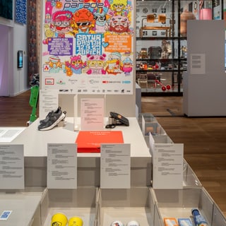 Museumsausstellung mit farbigem Poster und Gegenständen in Vitrinen.
