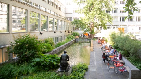 Innenhof mit Teich, Statue, Tischen und Stühlen unter Bäumen.