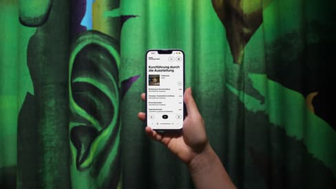 Hand hält ein Smartphone vor einem grünen Vorhang mit einem grossen Ohr.