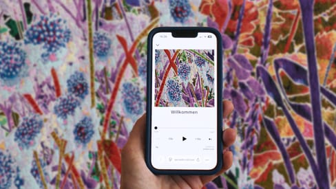 Hand hält Smartphone vor farbigem Blumengemälde.
