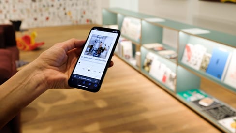Hand hält Smartphone in Kunstgalerie mit Bücherauslage.