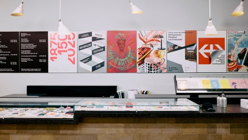 Innenansicht einer Ausstellung mit Plakaten an der Wand und einem Tresen im Vordergrund.