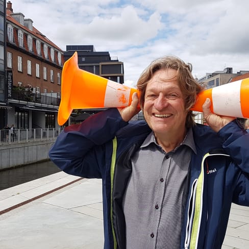 Mann hält orangefarbene Verkehrshütchen an Ohren, Strasse im Hintergrund.