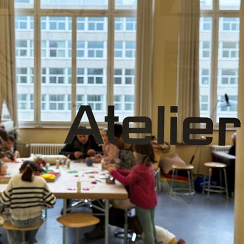 Blick durch Glastür mit Aufschrift 'Atelier' auf Menschen am Tisch kreativ arbeitend.