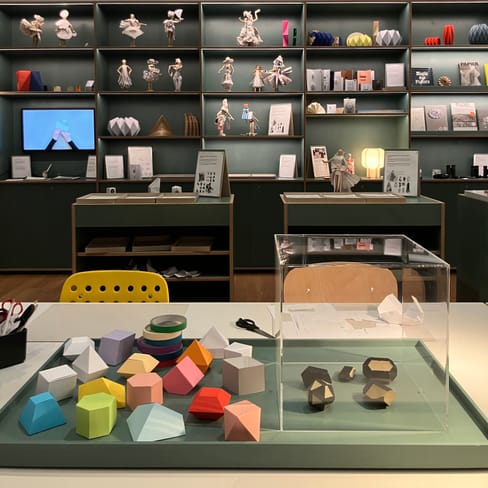 Tisch mit bunten geometrischen Objekten und Regalen mit Dekorationen im Hintergrund.