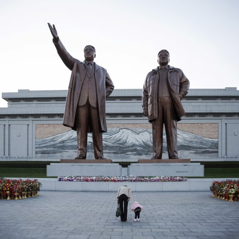 Zwei grosse Bronzestatuen auf einem Platz mit Blumen und Gebäudefassade im Hintergrund.