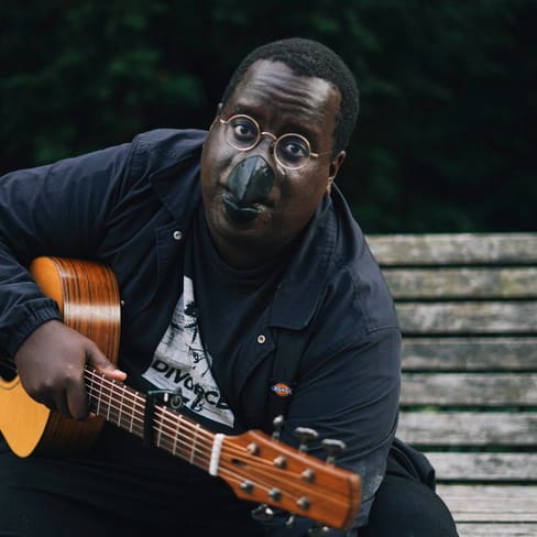 Person mit Gitarre und schwarzer Gesichtsmaske auf Bank sitzend.