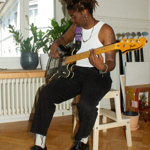 Person spielt E-Bass auf einem Stuhl sitzend in einem Zimmer mit Pflanzen im Hintergrund.