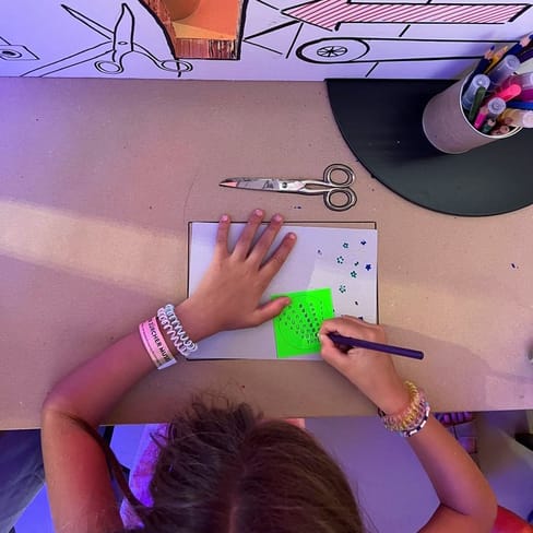 Kind schreibt auf grünem Papier an einem Schreibtisch mit Stiften.