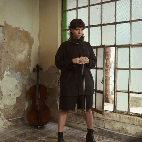 Person in schwarzer Kleidung in einem alten Raum mit Cello und grossen Fenstern.