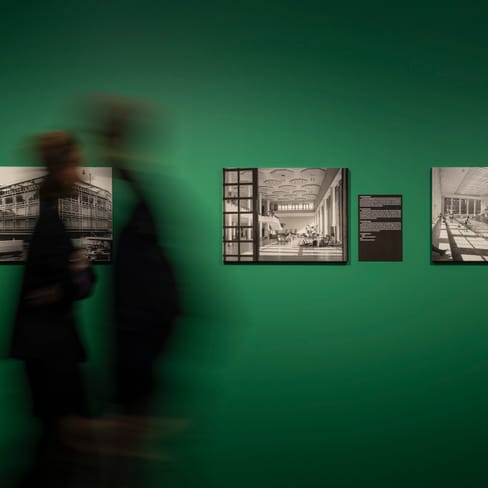 Zwei unscharfe Personen vor grüner Wand mit schwarz-weissen Fotos.
