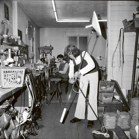 Person in Werkstatt, arbeitet an Lampe mit Werkzeugen im Hintergrund.