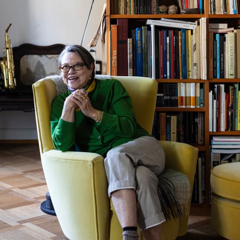 Stefi Talman sitzt auf gelbem Sessel vor Bücherregal in ihrer Wohnung.