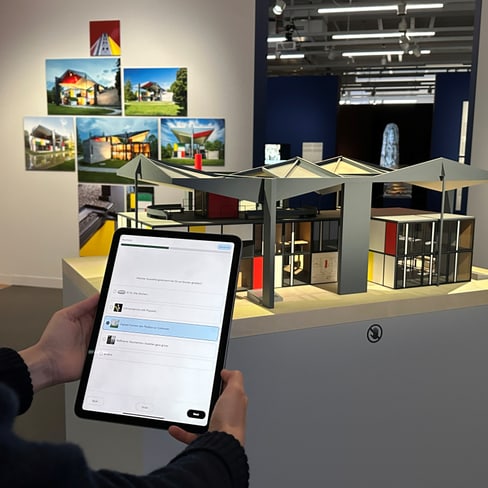 Person hält Tablet vor Architekturmodell und Fotos an der Wand.