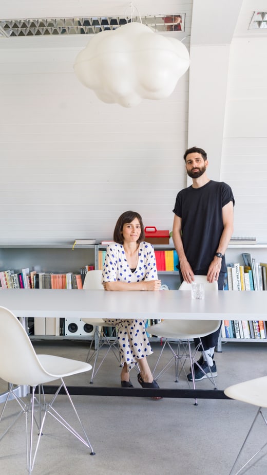 Zwei Personen an einem Tisch in modernem Raum mit Bücherregal und Wolkenlampe.