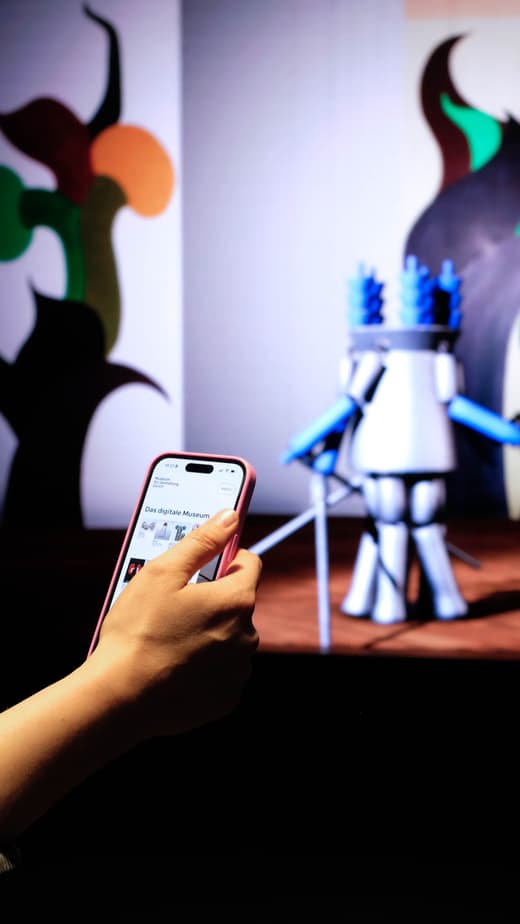 Person hält Smartphone vor einem Kunstwerk mit bunten Figuren.