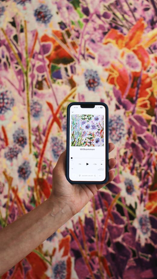 Hand hält Smartphone vor bunter Blumenwand.