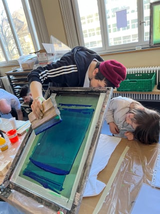 Person druckt mit Kind, Materialien auf Tisch in sonnigem Raum.