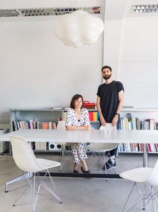Zwei Personen an einem Tisch in modernem Raum mit Bücherregal und Wolkenlampe.