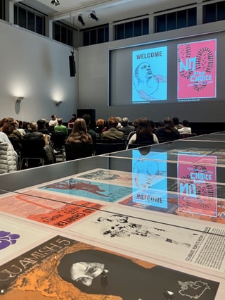 Museumsausstellung mit Plakaten und Vortragspräsentation, Publikum sitzt zuhörend.