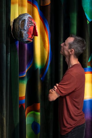 Person in einem roten Hemd schaut auf eine bunte abstrakte Maske an der Wand.