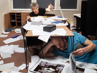 Zwei Personen unter und über Tisch mit Papieren und Büchern verstreut.