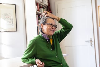 Ältere Frau mit Brille in grünem Pullover, streicht Haar zurück, sitzt lächelnd drinnen.