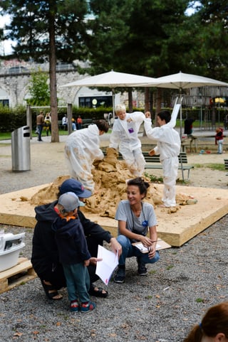 Kinder in Schutzanzügen basteln mit Lehm im Park. Eine Leitungsperson sitzt davor und erklärt das Projekt zwei einem Vater mit Sohn.