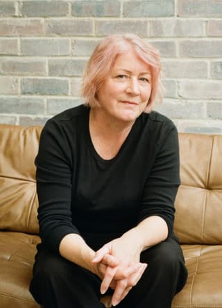 Frau mit rosa Haaren sitzt auf braunem Sofa vor Ziegelwand.