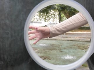 Hand, die durch einen runden Spiegel  gezeigt wird, mit Brunnen im Hintergrund.