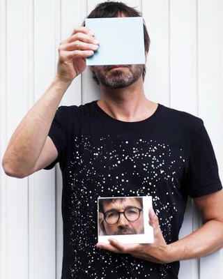 Person hält Foto vor Gesicht, verdeckt teilweise mit Buch, trägt schwarzes T-Shirt mit Muster.