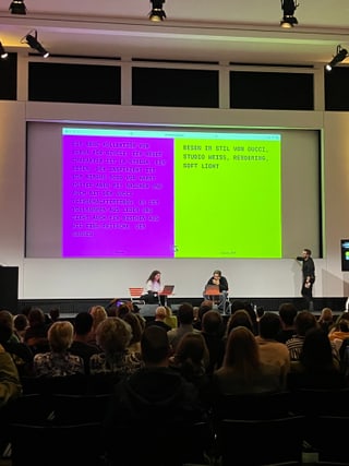 Präsentation vor Publikum mit zwei Sprechern auf Bühne und Textprojektion auf Leinwand.