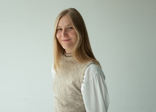Person mit langem blondem Haar lächelt leicht vor neutralem Hintergrund.
