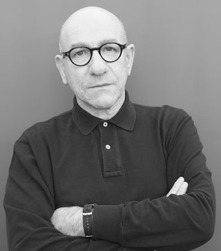 Mann mit Brille und verschränkten Armen vor neutralem Hintergrund.