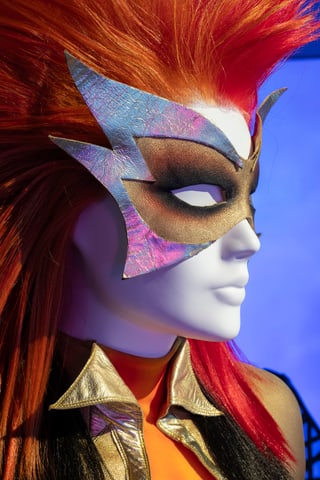 Mannequin mit rotem Haar und auffälliger Maske vor blauem Hintergrund.